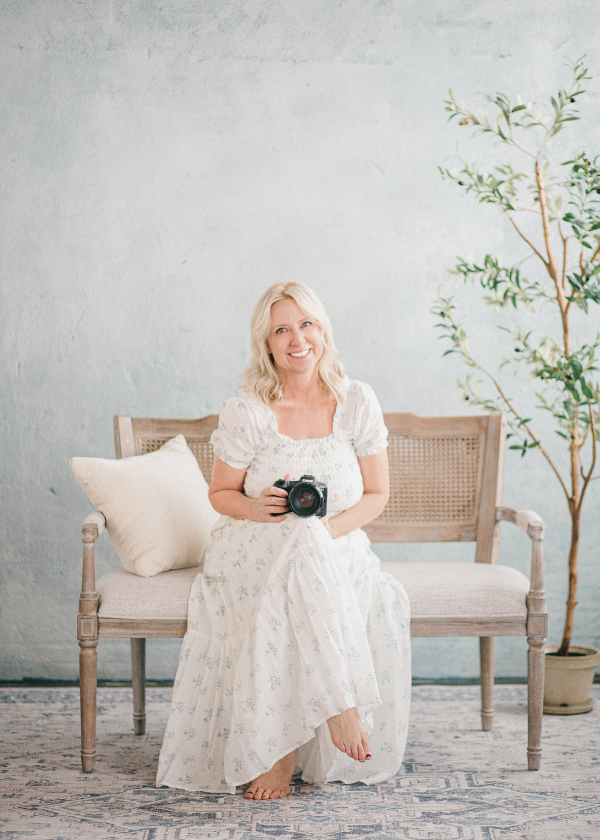 Woman in a white dress sits on a bench, holding a camera, against a light blue background with greenery.