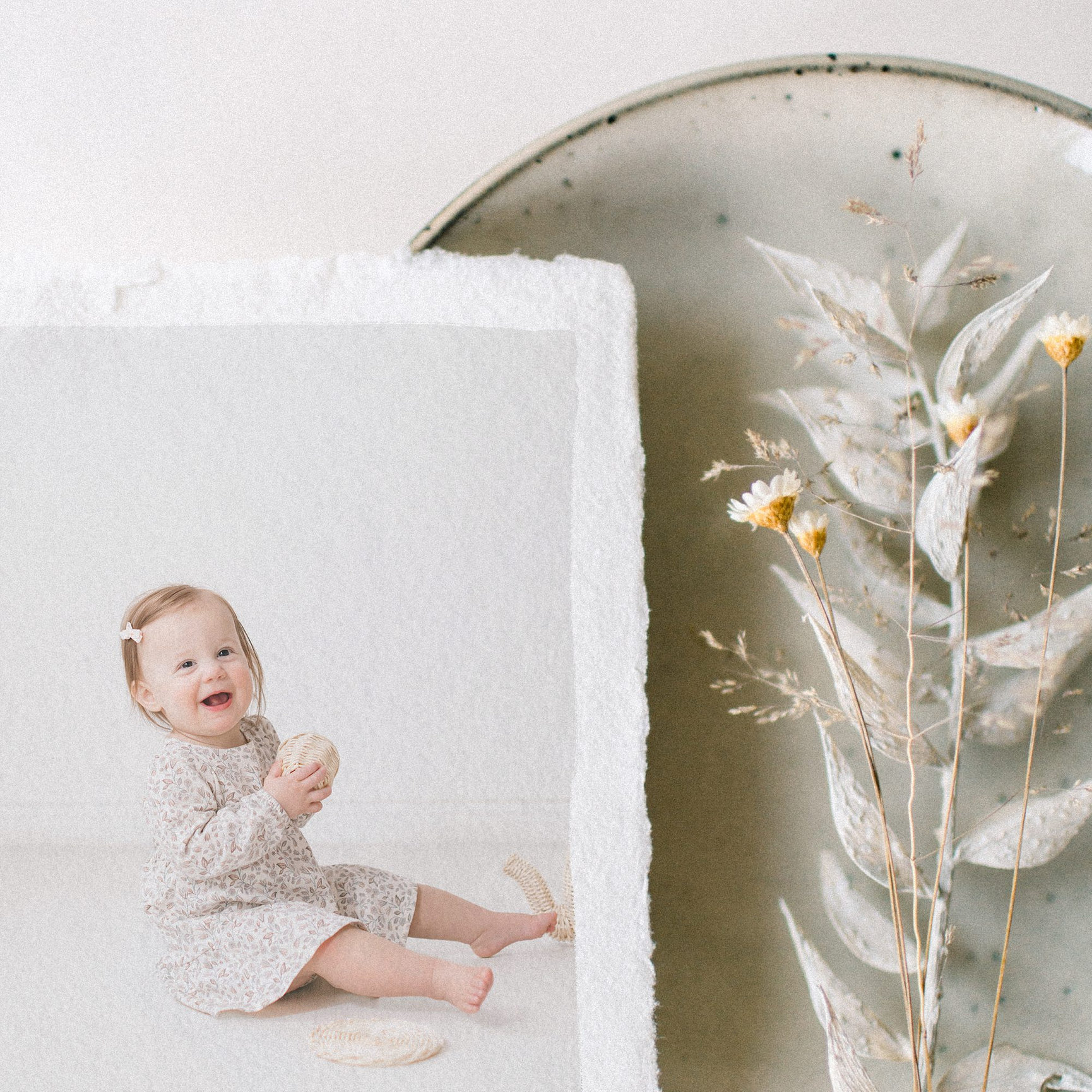 A smiling toddler in a light dress holds a snack, with a floral arrangement nearby. Soft, neutral colors dominate the scene.