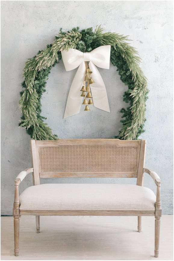 A minimalist scene featuring a green wreath with a bow and bells above a light-colored wooden chair.