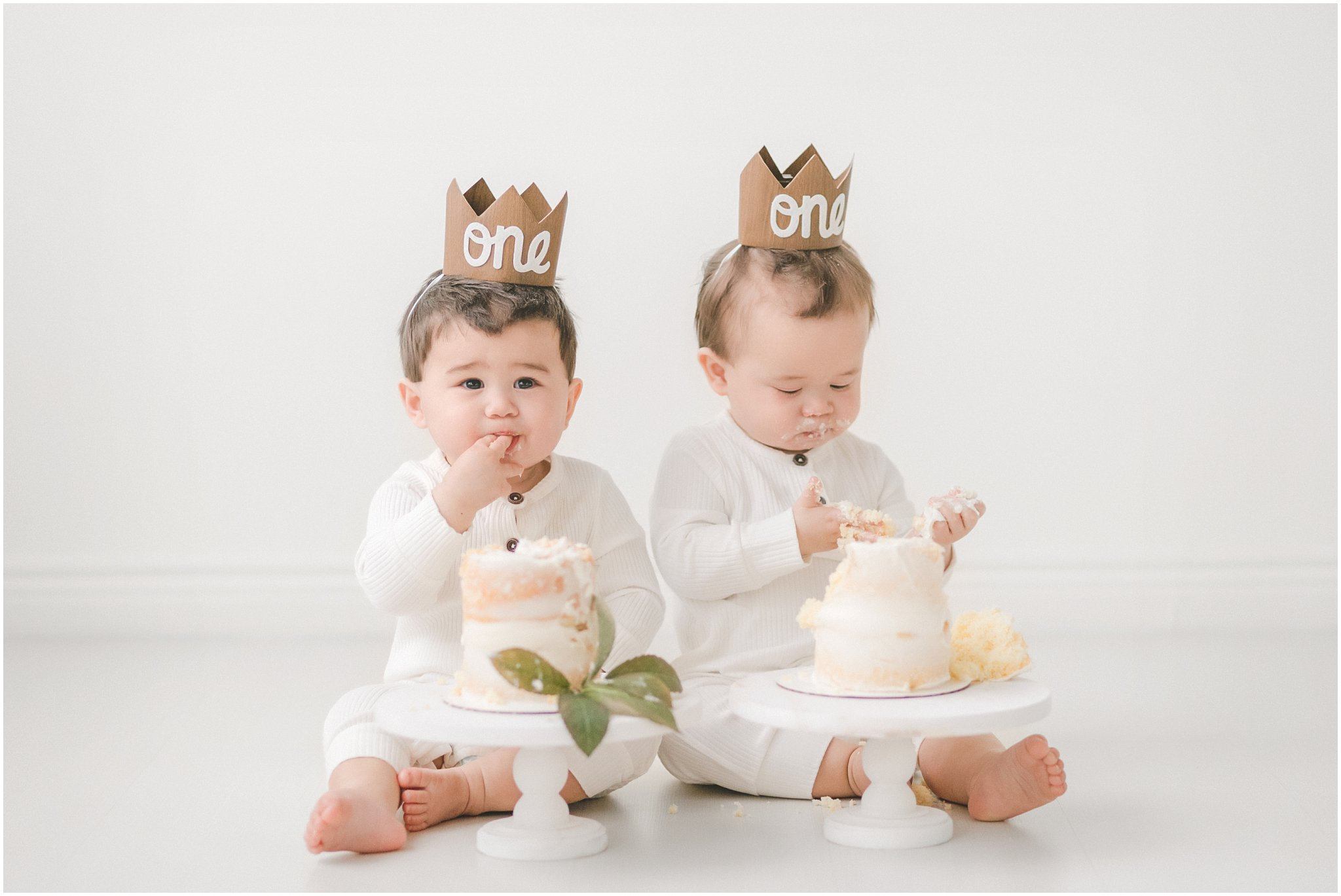 Two toddlers wearing crown hats sit on a white background, enjoying small birthday cakes on pedestals.