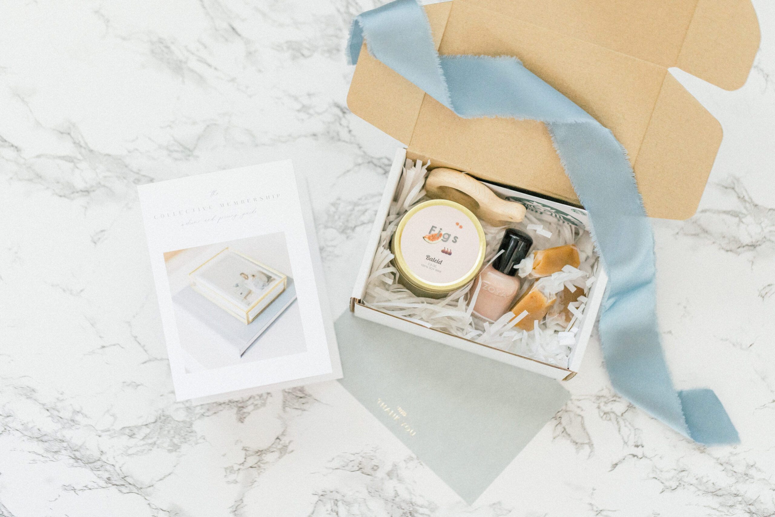 Gift box with a candle, brush, and ribbon on a marble surface, alongside a book.