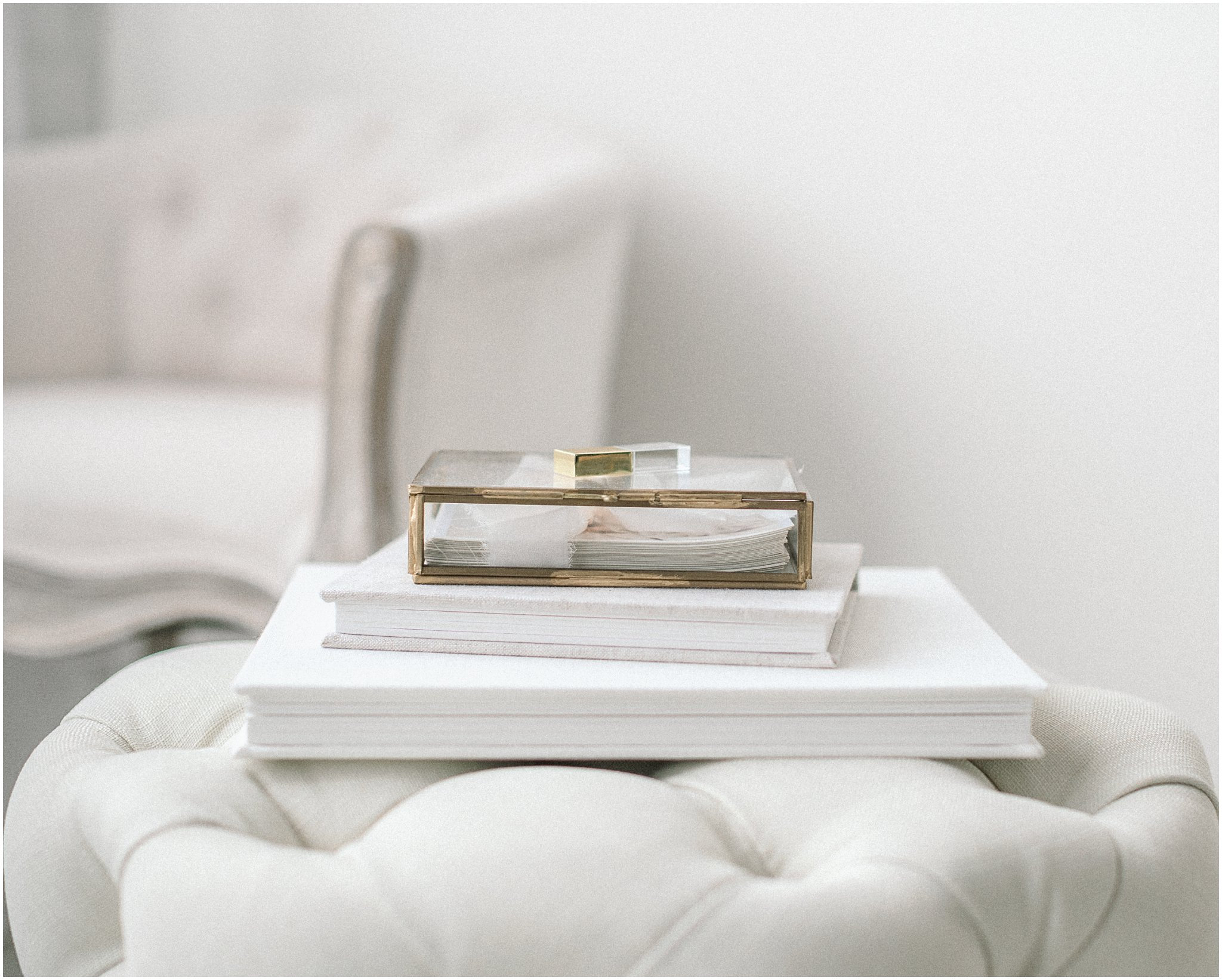Glass box with books on a white tufted ottoman, blurred chair in background.