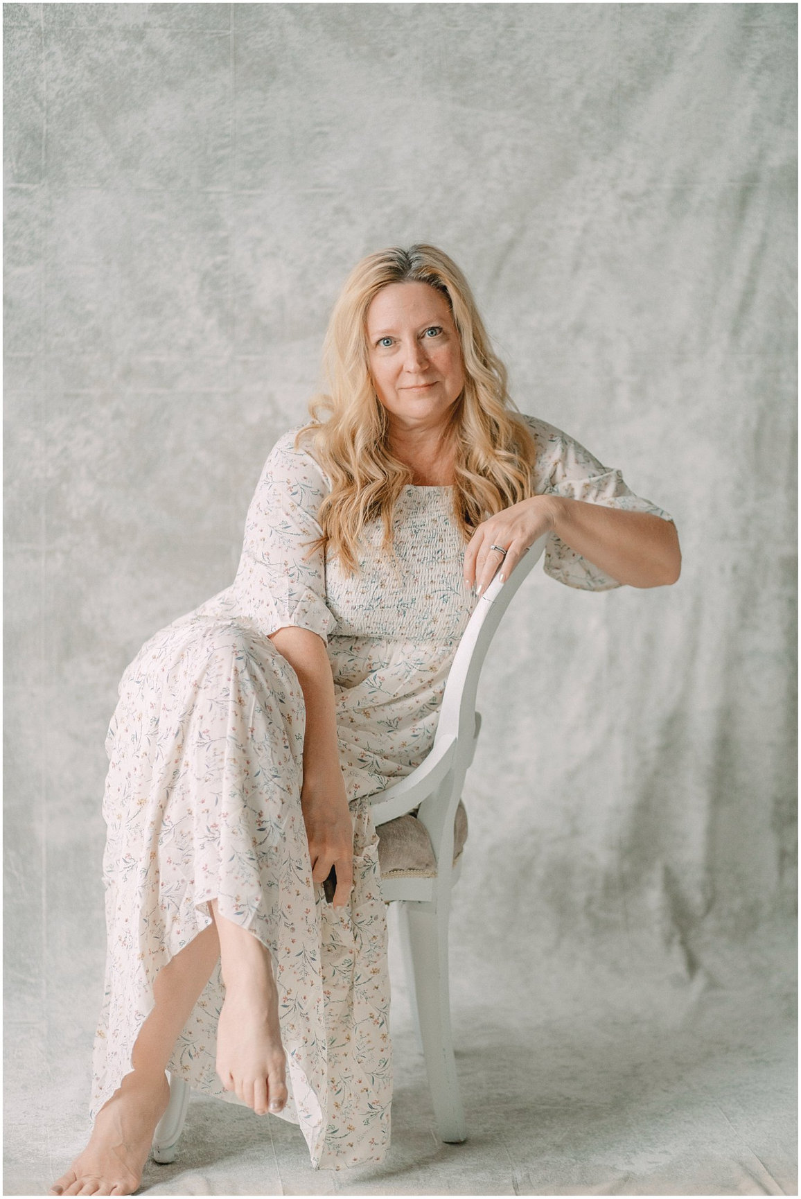 Woman in a light floral dress sitting on a white chair against a textured light gray background.