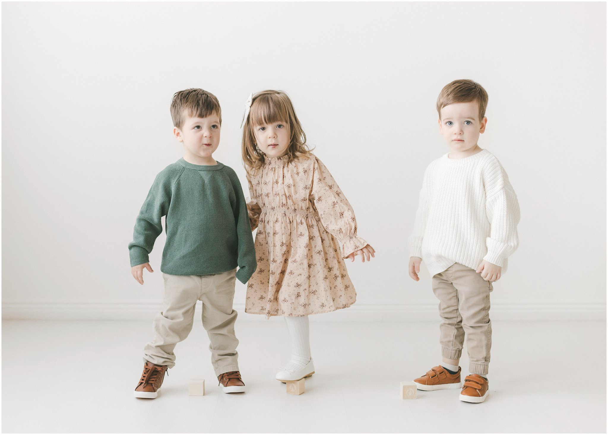 Three children standing together, one girl in a dress and two boys in sweaters and pants, against a white background.