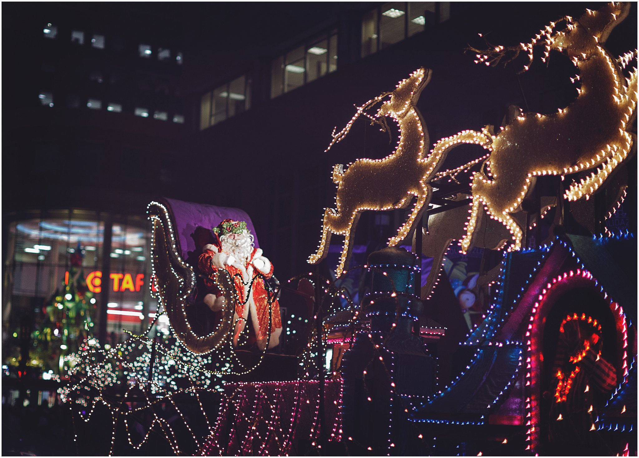 A festive holiday parade float with illuminated reindeer and Santa Claus in a city at night.