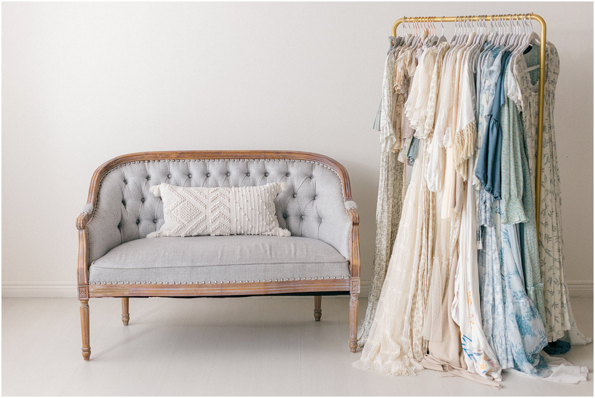 Wooden bench with a cushion next to a clothing rack filled with beige and blue dresses.