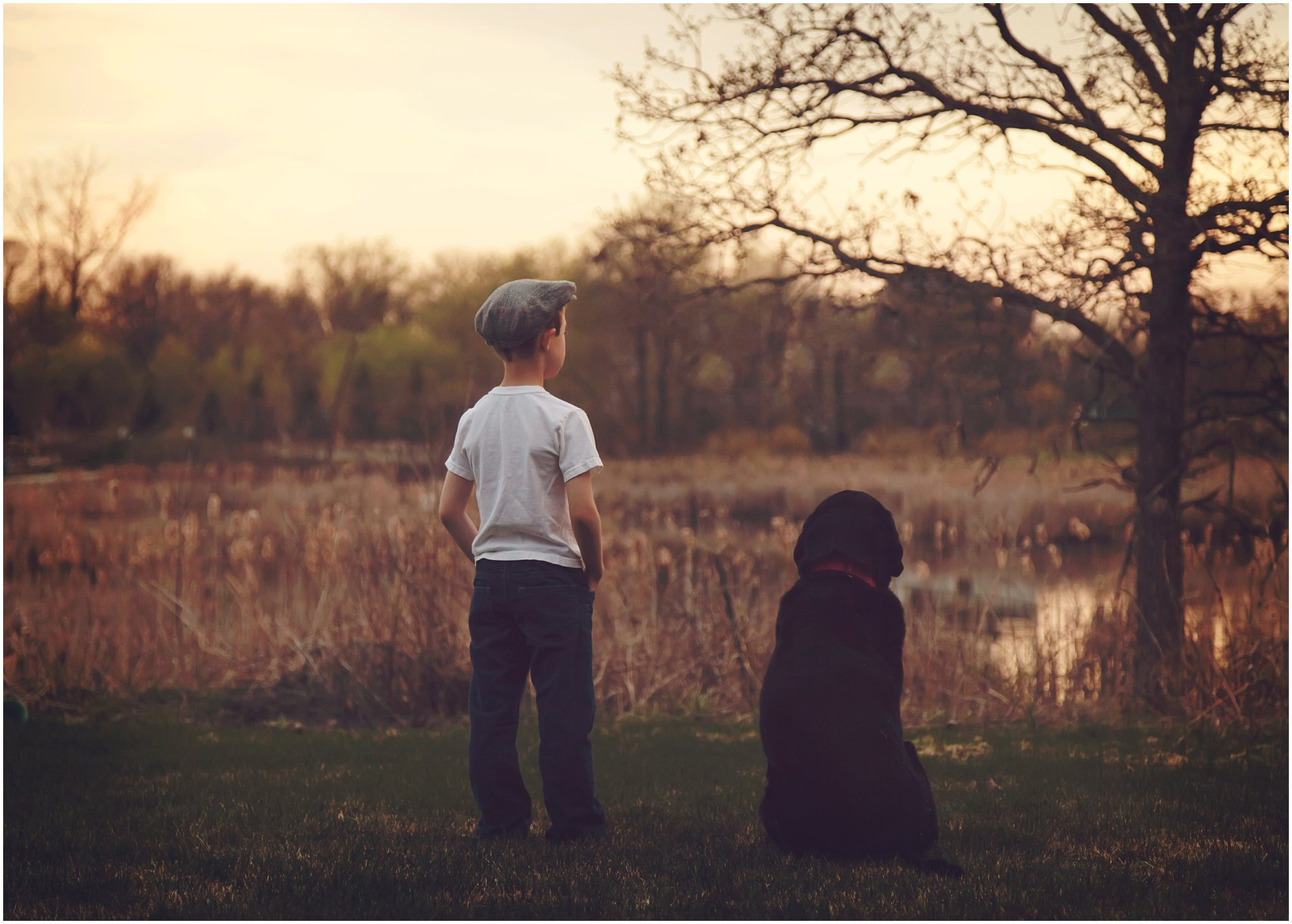 Child and dog standing by a lake with trees in the background at sunset.