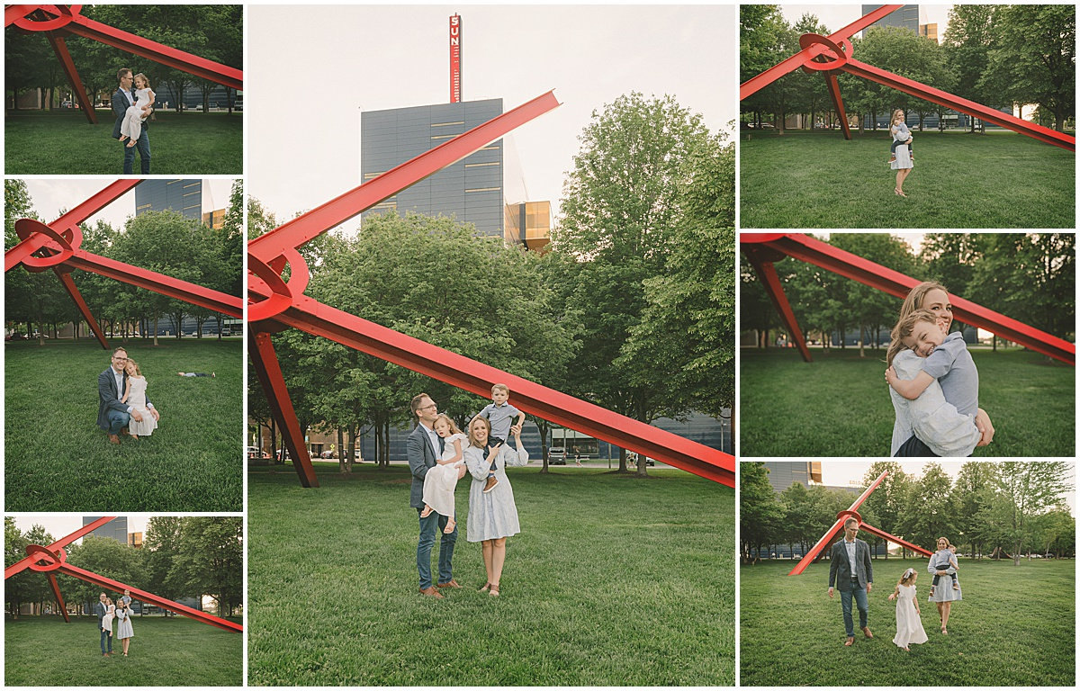 A couple poses under a large red sculpture in a park, with trees and a tall building in the background.