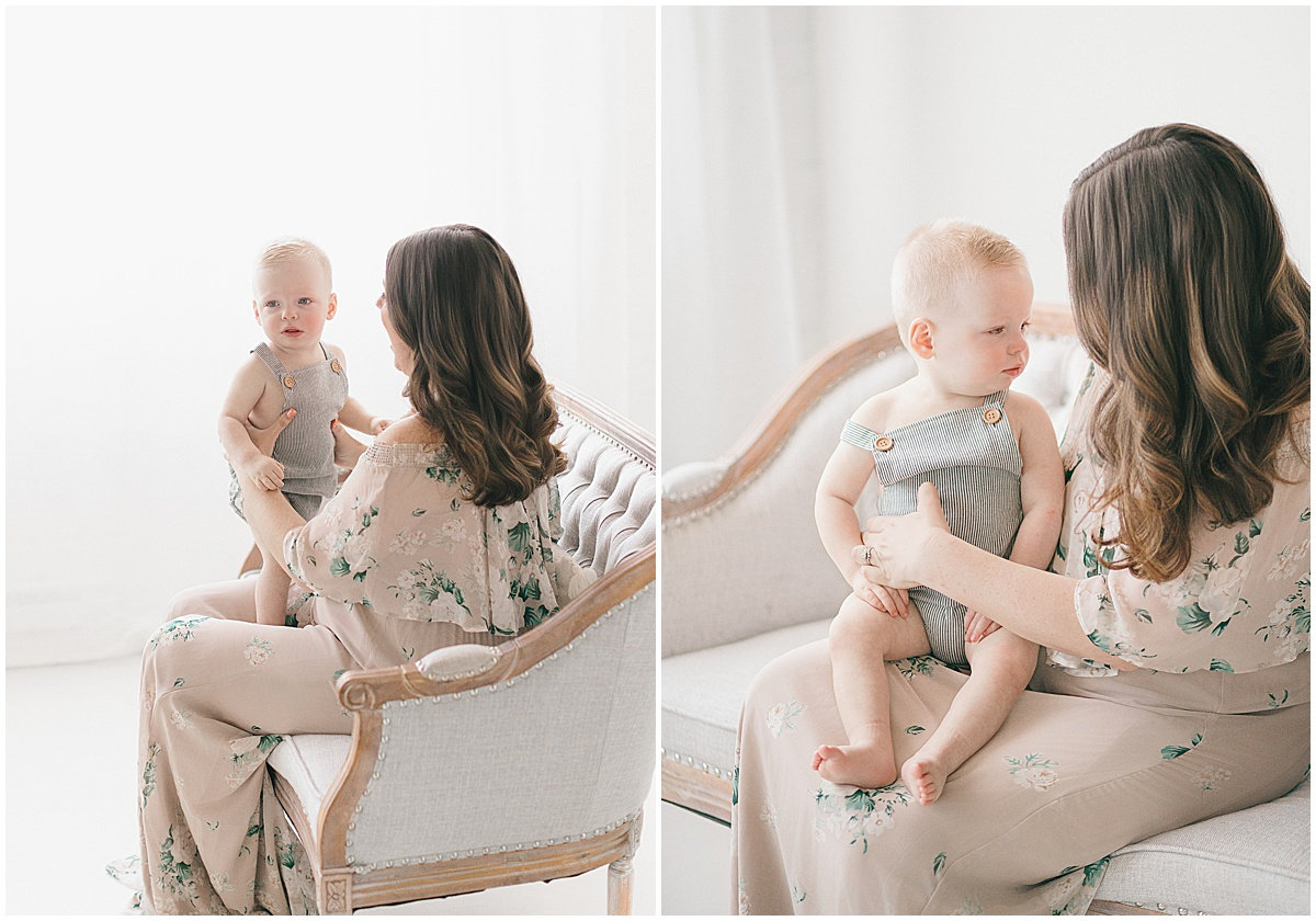 A woman in a floral dress sits on a sofa holding a smiling baby, both in a bright, softly lit room.