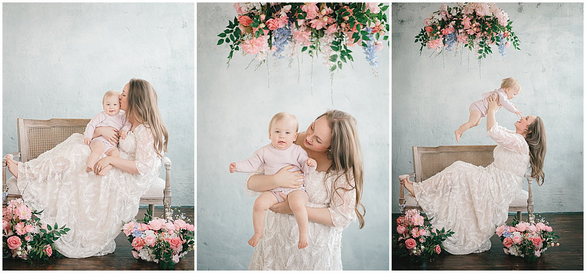 A mother in a white dress holds and plays with her baby, surrounded by pink flowers and green foliage in a light, airy setting.