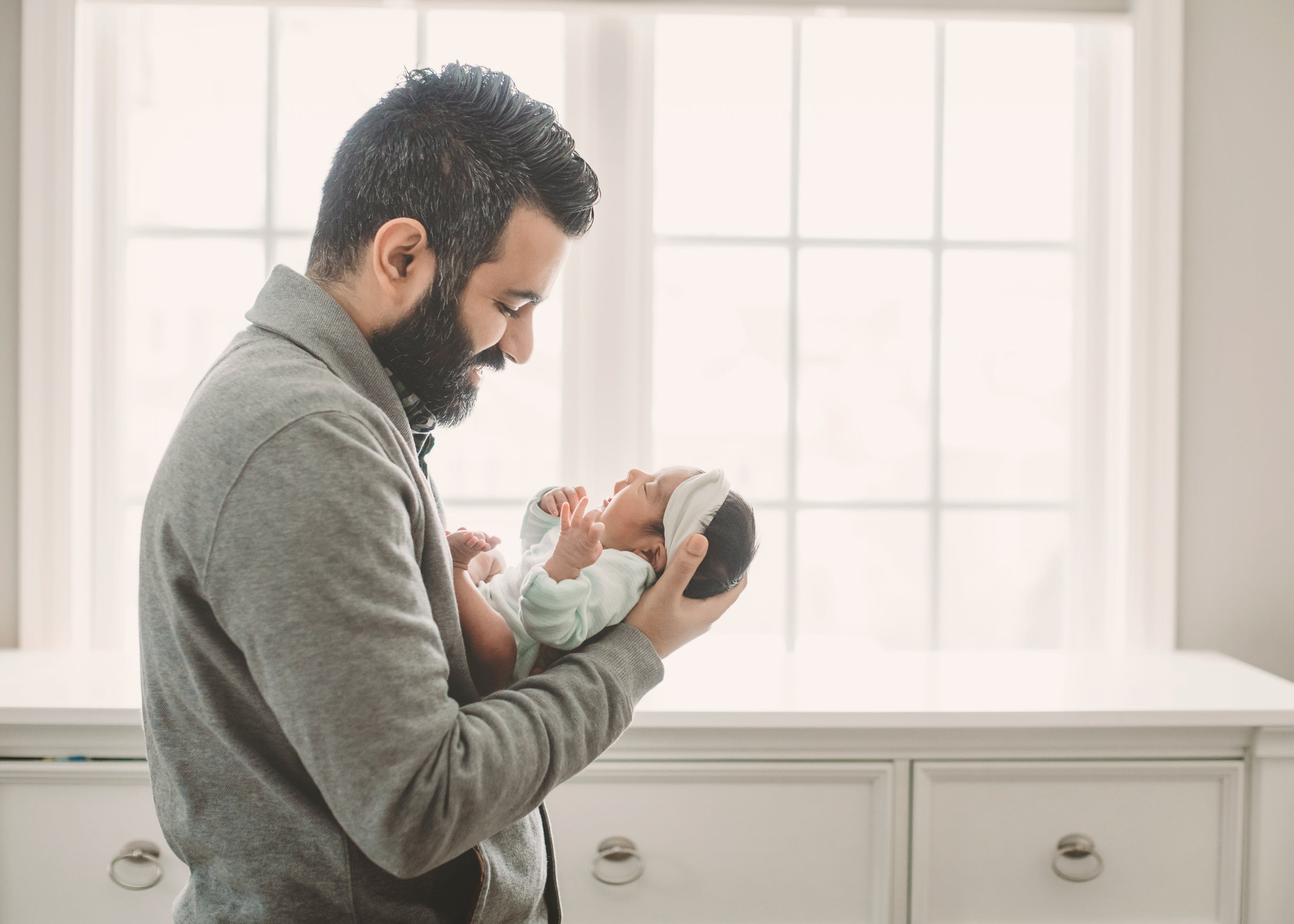 A man gently cradling a newborn in front of a sunlit window.
