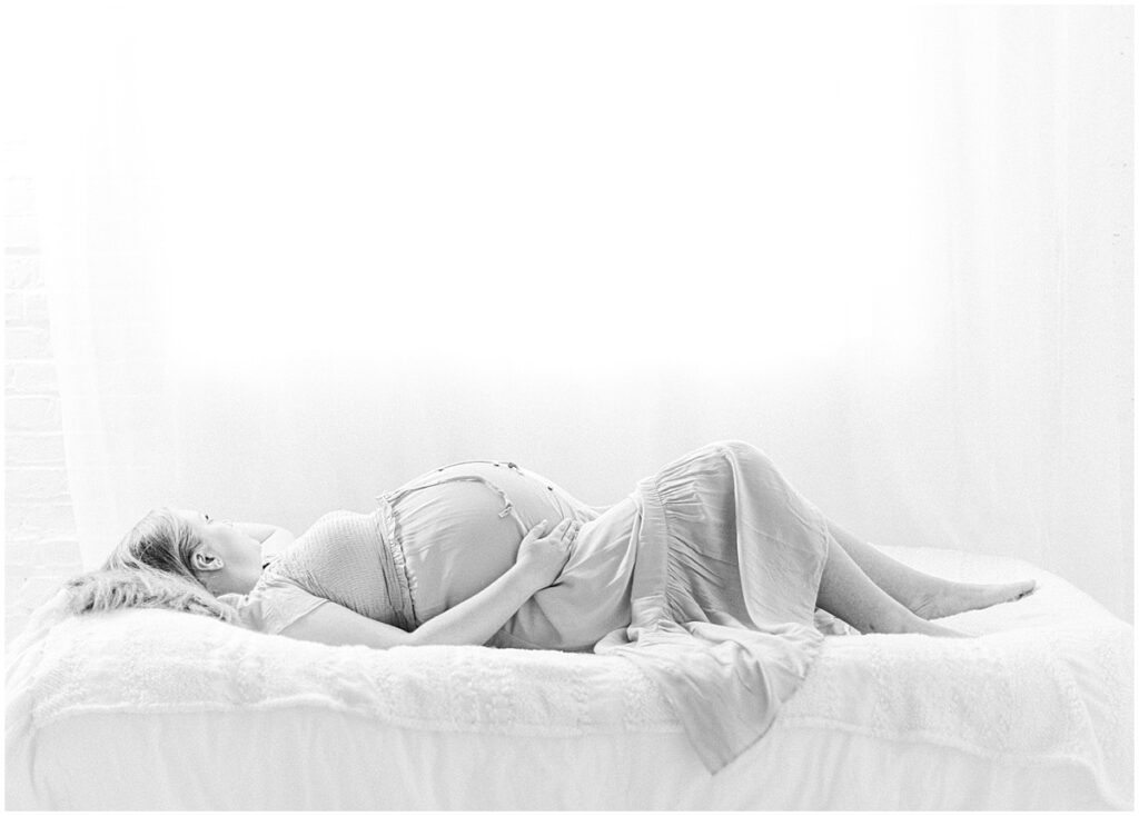 Black and white maternity portrait of a pregnant woman reclining on a bed with one hand resting near her belly