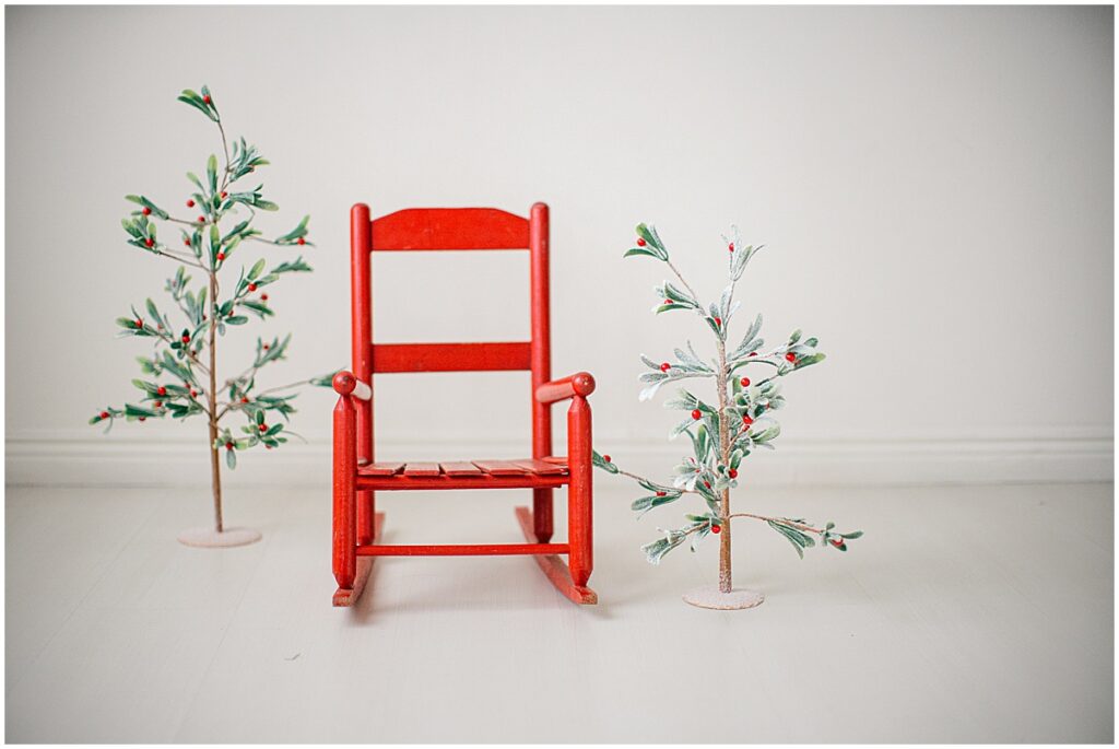 Red wooden rocking chair between two small holiday trees for a simple Christmas mini session setup.
