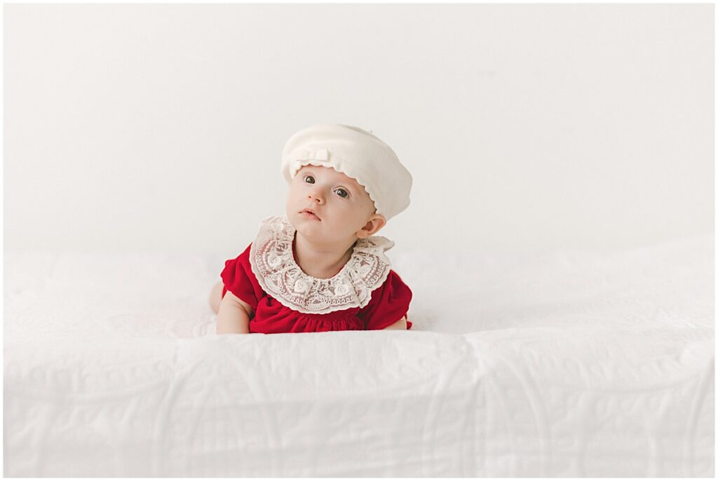 Baby in a red holiday outfit and white bonnet photographed on a simple white studio set.